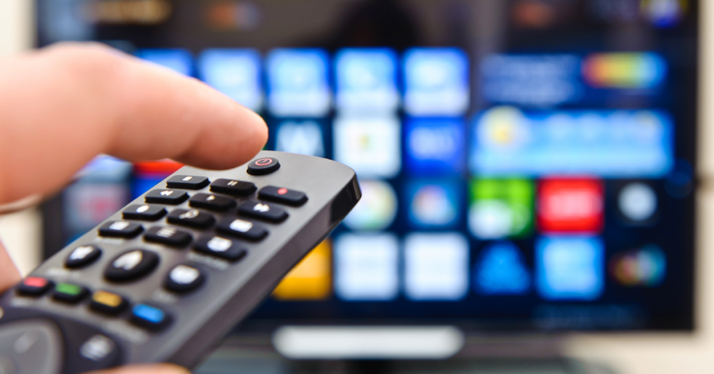 A close-up image of a hand holding a remote control, pointed towards a television screen. The TV screen in the background is slightly blurred and displays a variety of colorful app icons, indicating a smart TV interface.