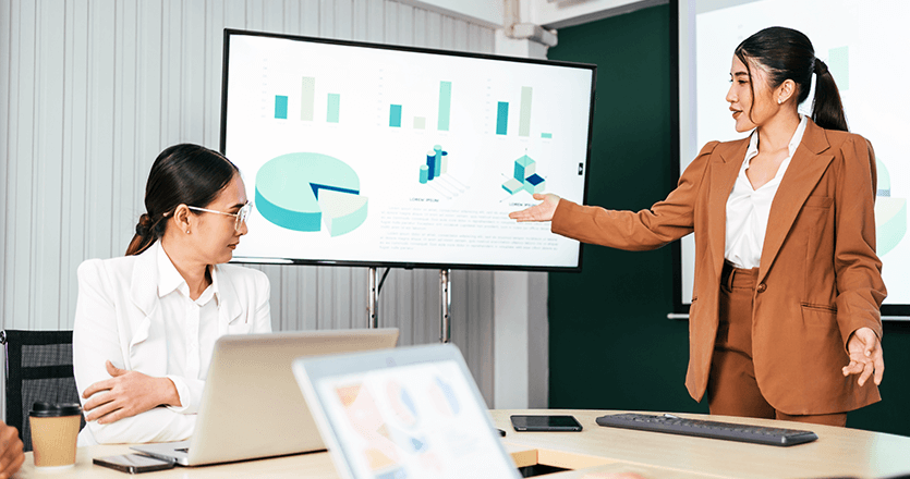 An image of two employees in an office looking at a digital screen. In the screen there are displayed some graphs. 