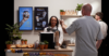 Café set up, with digital signage screens and a customer pointing at them. A cashier in front of the screens smiling.