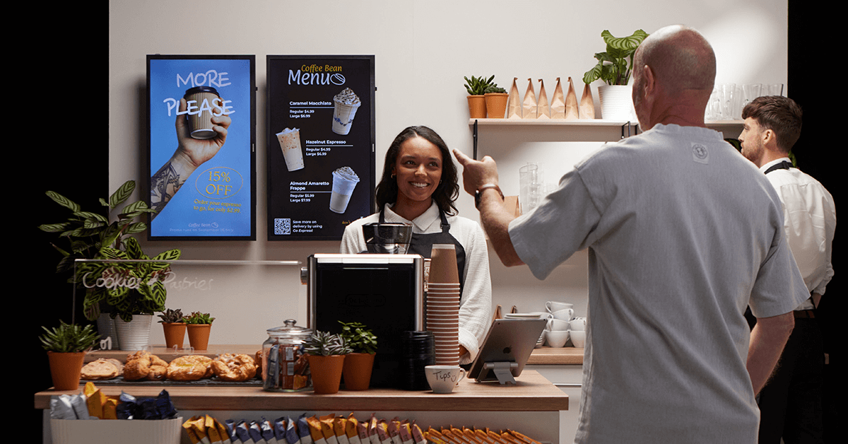 Café set up, with digital signage screens and a customer pointing at them. A cashier in front of the screens smiling.